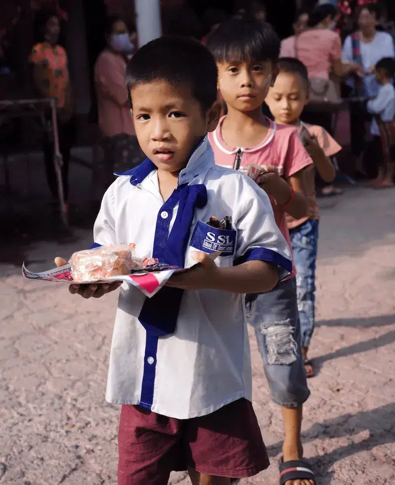 A young boy in a school uniform carrying a plate of food, with other children lined up behind him