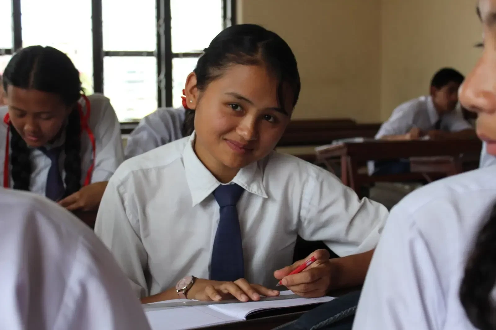 A smiling student in a white shirt and navy tie looks up from her exercise book in a classroom